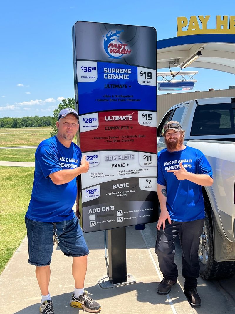 Two Fast Wash crew members in blue Ask Me About Memberships shirts pointing at the wash menu board
